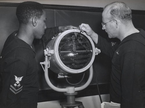 "The functions of the various parts of a 'blinker' are explained in detail by Signalman First Class Ernest V. Alderman, USNR, to Signalman Second Class Julius Holmes, who will be operating one aboard a DE [destroyer escort]. 