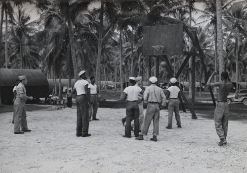 "'Recreation for Negro Seaman in South Pacific' - Full expression is allowed for recreational activities for Negro seamen stationed on Espiritu Santo in the New Hebrides. In their off-duty hours they play baseball, basketball, and other outdoor and indoor sports. Though far from home, their combined energies have made of the tropic base a palm-decorated 'home away from home' as they serve on the far-flung Pacific front. This photo shows one of the basketball teams in action."
