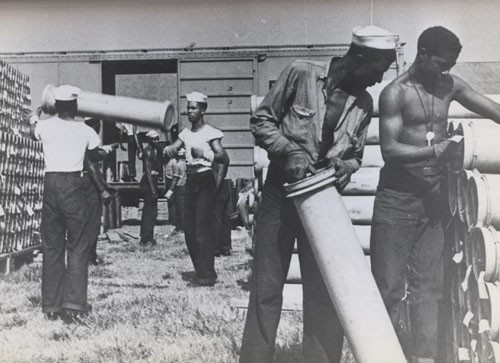 "'Negro enlisted men play ordnance 'Shell Game'.' - One of the unending tasks for enlisted men assigned to Naval Ordnance is the handling of empty cases in which shells are shipped. These colored Navy men at St. Juliens Creek Ammunition Depot in Virginia stack empties which held five-inch shells. The more than 6,000 Negro enlisted men in Navy Ordnance may be given any of the duties which fall to all enlisted men, ranging from manual tasks to hazardous assignments requiring great skill, such as handling live ammunition in a combat zone."