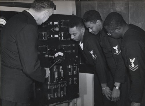 "Electrician's Mates (left to right) Nathaniel O. Dyson, Second Class; Richard Hubbard, Third Class; and John W. Reagan, Second Class, listen as Chief Electrician's Mate John E. Taylor, USN, (Ret.), explains the workings of the power system and distribution process they'll be working with when serving aboard a DE [destroyer escort]. 
