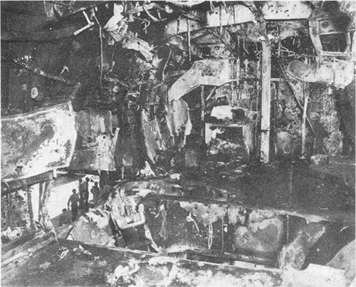 Photo E-15: Second hit. View of 5-inch Group III gun gallery looking forward showing damage in gallery deck and inboard bulkhead by explosion of bomb and burning of 5-inch ready-service ammunition.