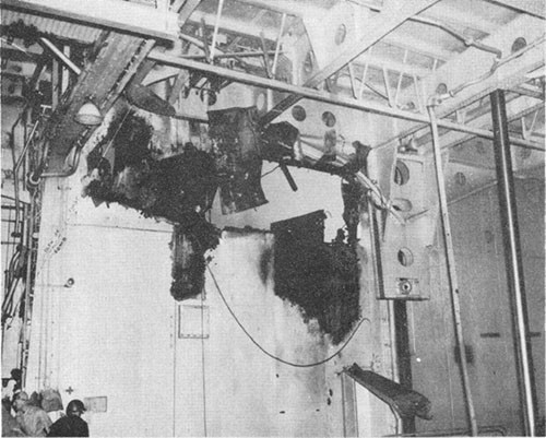 Photo E-16: Second hit. Looking outboard from No. 3 elevator trunk through hole blown in inboard bulkhead of Group III gun gallery by explosion of bomb and burning of 5-inch ready-service ammunition.