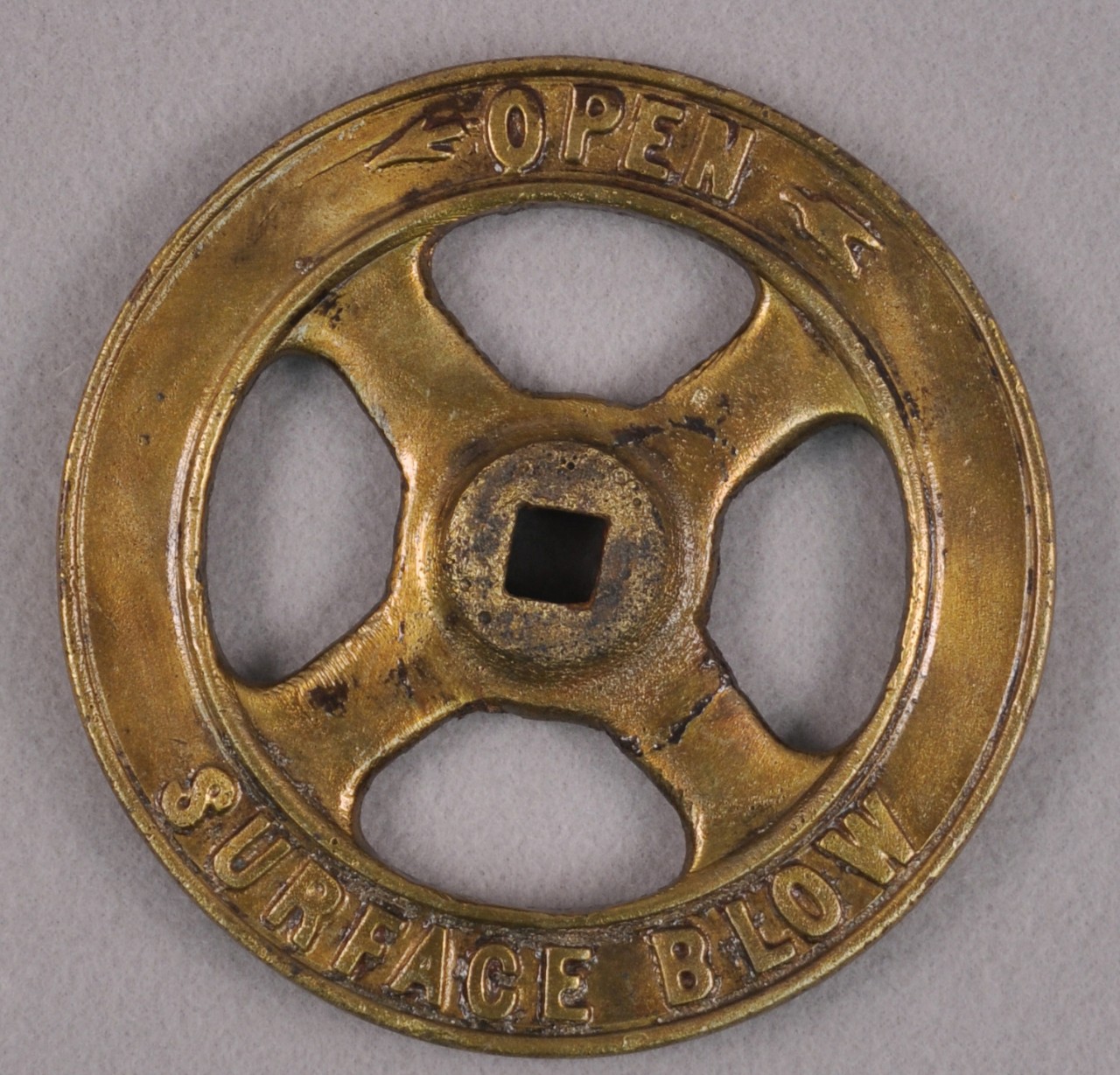 <p>A brass valve wheel with a raised circle in the middle. A square hole is cut in the middle of this circle. At the top of the valve are the words “OPEN” with an arrow point left and the words at the bottom are the words “SURFACE BELOW”.</p>
