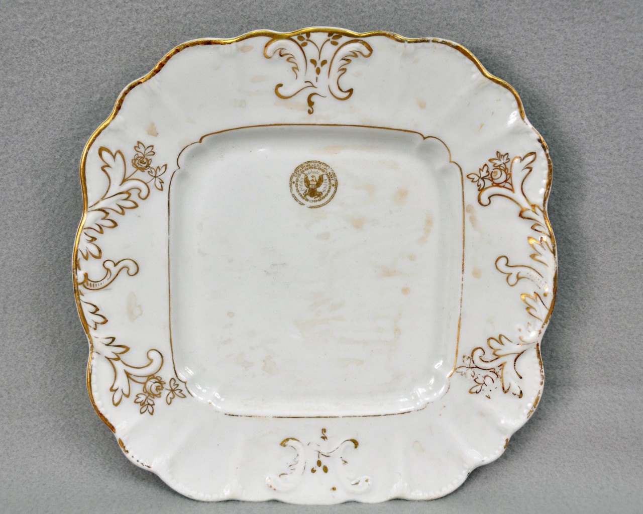 <p>A white, ceramic, square-shaped plate. It is decorated with gilded floral molding on the lip and has a gold eagle seal that says “Department of the Navy” on the top.</p>
