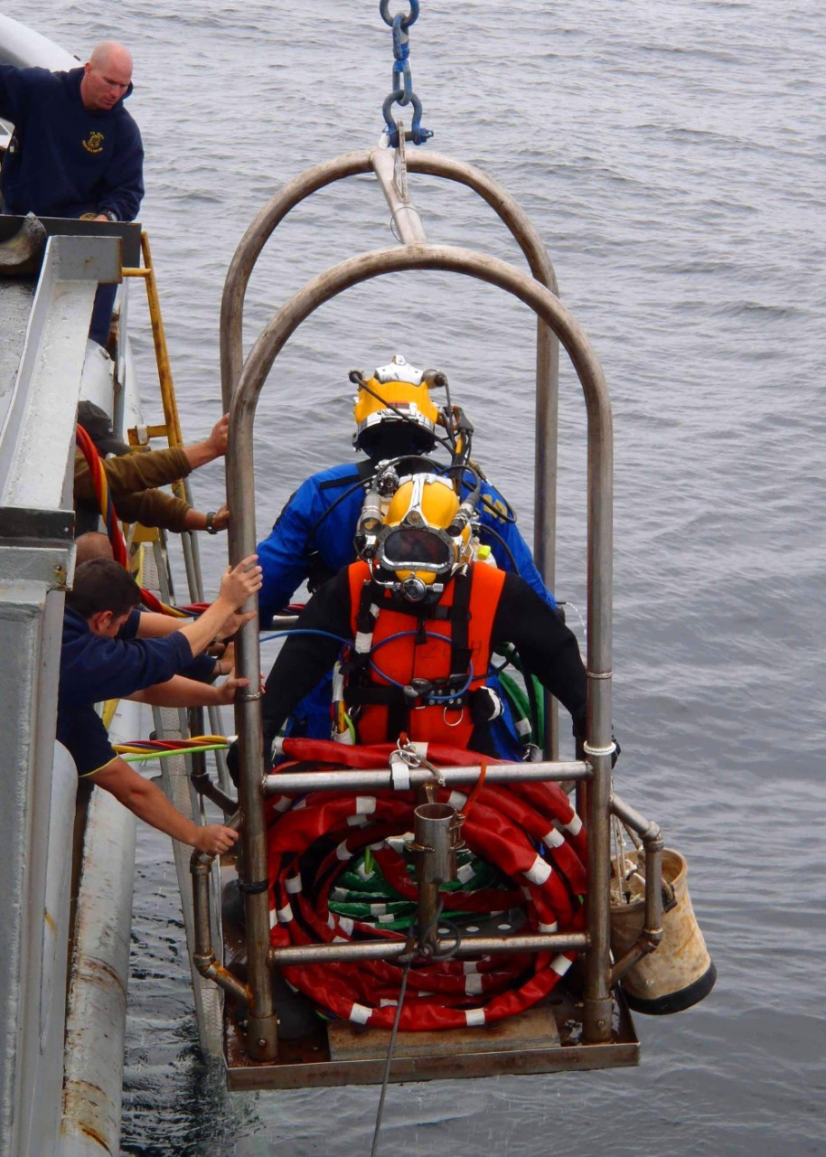 Two Navy divers wearing yellow helmets and blue dry suits on a mobile platform being lowered into the North Sea