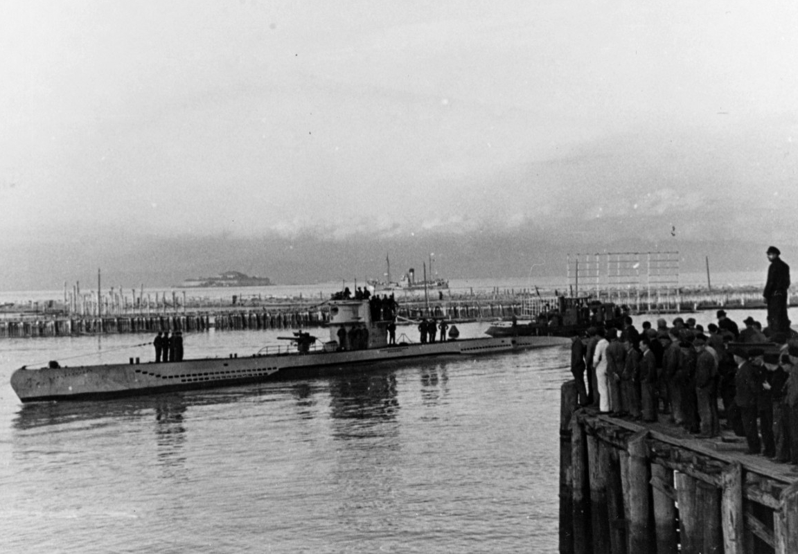 Black and white photograph of German submarine running at the surface with men standing on the deck., while people look on from shore.