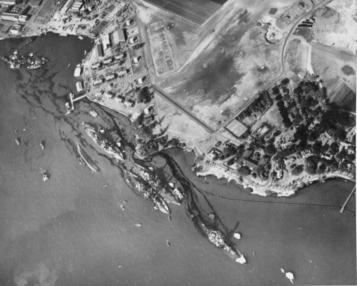 Black and white photograph looking down on the land and water of Pearl Harbor, with at six large damaged hulls leaking trails of oil.