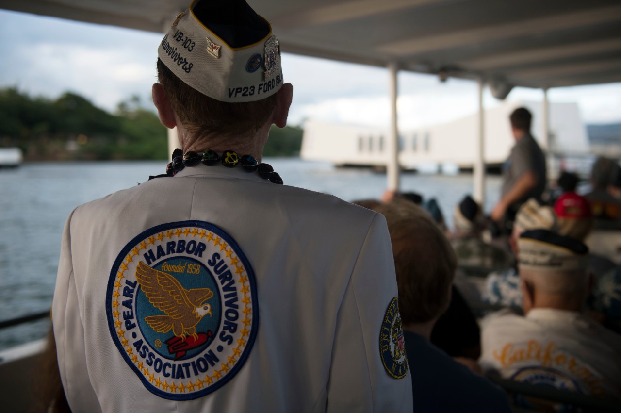 Color photo of a the back of a man wearing a jack with large patch that reads Pearl Harbor Survivors Association. The Arizona Memorial is in the background.