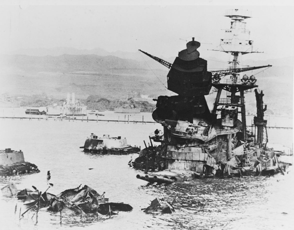 Black and white photo showing the superstructure of USS Arizona emerging from the water. The foremast is canted forward and the barrels of the three 14 inch guns of Turret 2 are elevated just above the water.