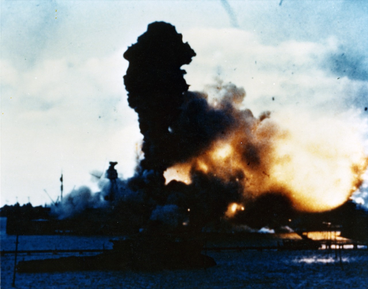 Color Photo of an explosion engulfing battleship USS Arizona (BB-39) with debris flying in the air.
