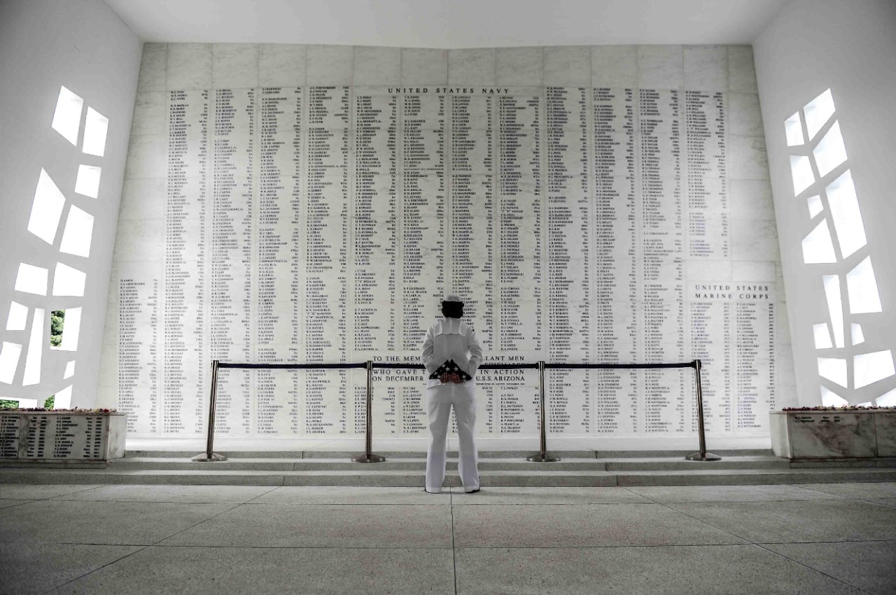 Color photo from behind a Navy Sailor in enlisted service dress white uniform centered in front of a large stone wall with 12 full columns and 2 half columns of inscribed names.