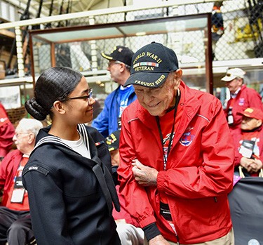 Link goes to Ceremonies and Events.  Image shows a Naval History and Heritage Command Sailor interacting with a Navy World War II veteran during the cutting of the cake on a Navy Birthday.   