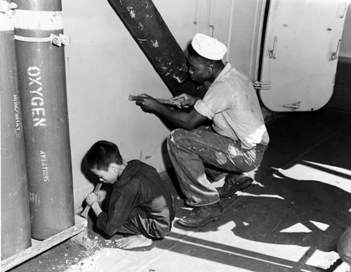 80-G-644528  Operation "Passage to Freedom", 1954  A young Vietnamese refugee lends a helping hand to a sailor chipping paint on board USS Bayfield (APA-33), while enroute from Haiphong to Saigon, Indochina, 7 September 1954.  Note aviators' oxygen bottle at left.  U.S. Navy Photograph, now in the collections of the National Archives.
