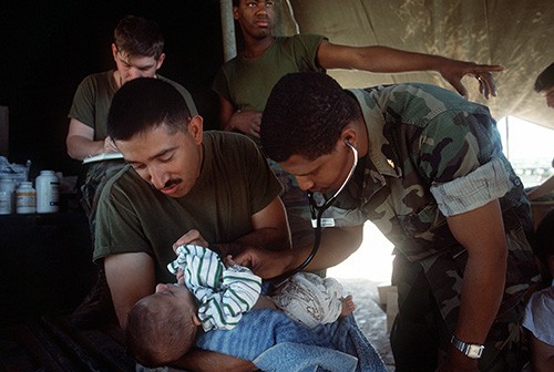 330-CFD-DN-ST-91-09848:   Operation Provide Comfort.   Hospital Corpsman First Class Charlie Alva, left, holds a child as Lieutenant Ronald Buckley listens to the child’s heart in a medical tent set up on the outskirts of the city.  Alva and Buckley are U.S. Navy medical personnel attached to Marine Expeditionary Unit (MEU) Service Support Group 24 (MSSG-24), which is providing services for Kurdish refugees.  Photographed April 27, 1991 by PH2 Savage.   Official U.S. Navy photograph, now in the collections of the National Archives.   