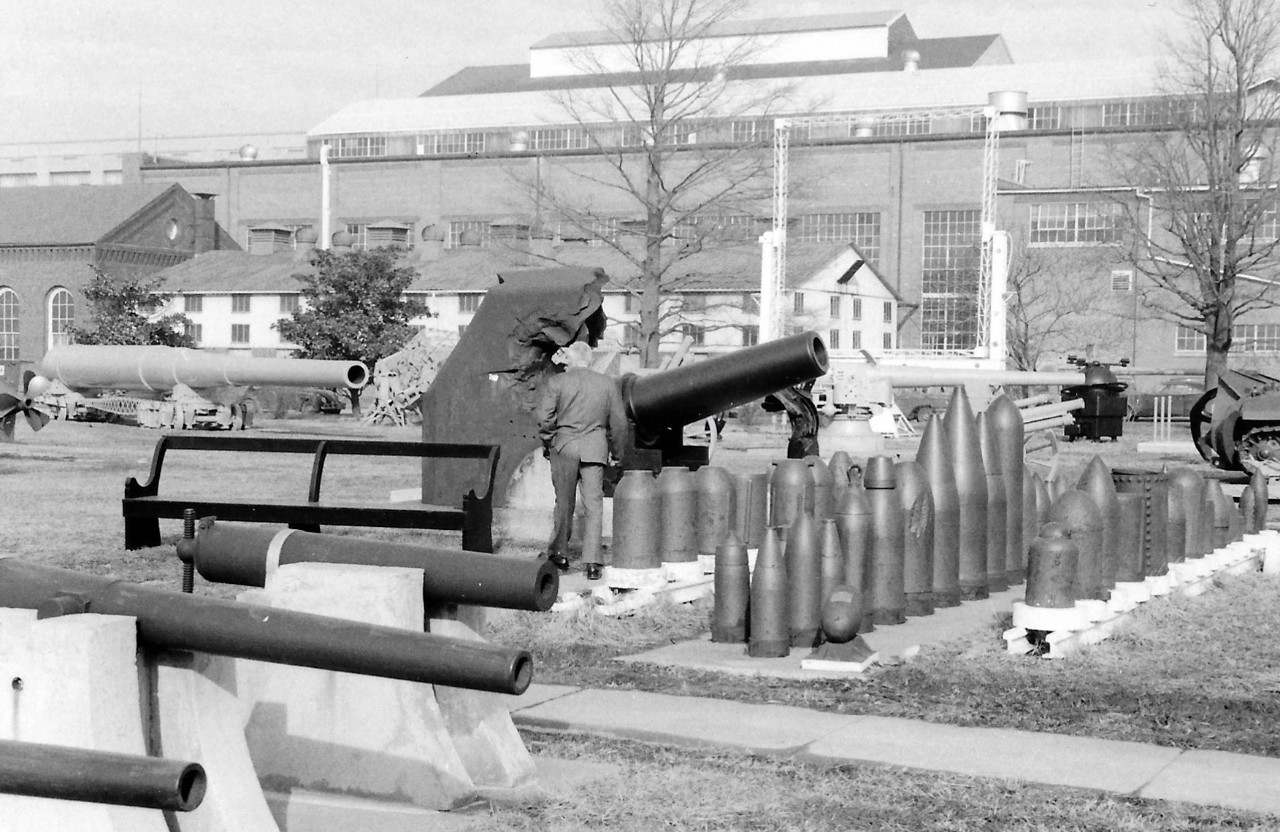 NMUSN-159:  Willard Park, Washington Navy Yard, Washington, D.C. 1970s.   View looking north.    To the right:  Japanese shells from World War II.  Center:  Yamato turret plate just beyond gentleman; with the 12” gun and German 5” naval gun showing beyond the Japanese shells.   Note the 16” gun to the mid-center left.   National Museum of the U.S. Navy Photograph Collection. 
