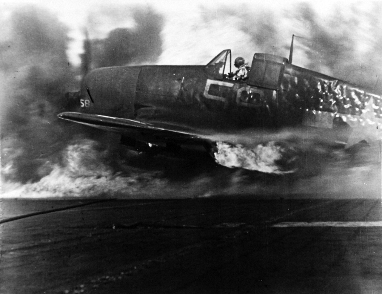 80-G-49697: Japanese Kamikaze Attack, June 1945. View of wounded pilot making a landing with his burning plane which was hit by a kamikaze.  The plane is aflame, smoke rolls around it as firefighters not seen in this picture converge on it.    Released June 28, 1945.   Official U.S. Navy photograph, now in the collections of the National Archives.  