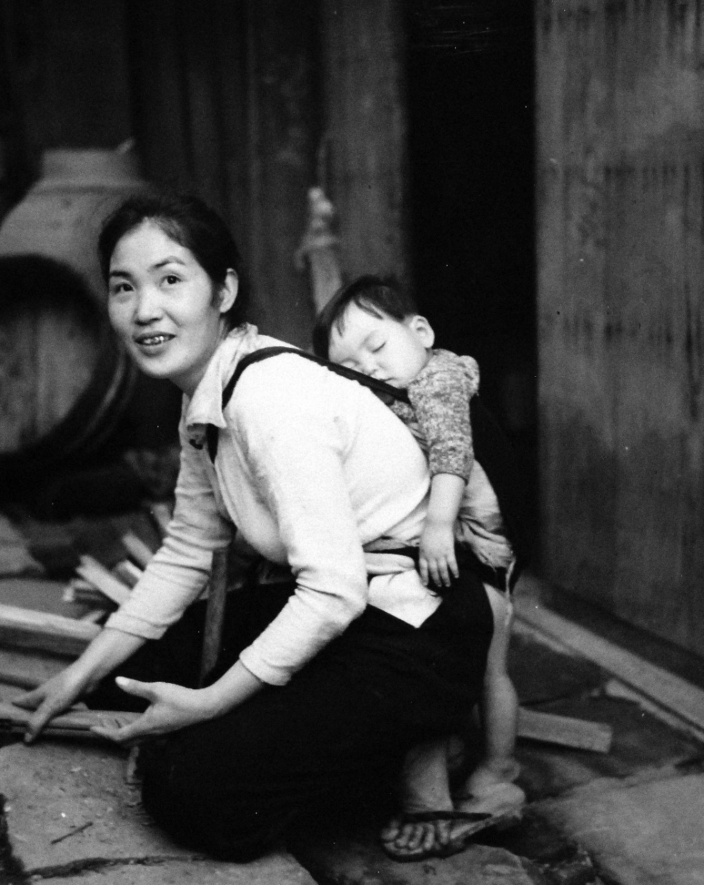 80-G-264848:   Occupation of Japan, 1945.    Scene at Sasebo, Kyushu, Japan, shows mother working with sleeping child on her back.  Photographed by crewmember of USS Chenango (CVE-28), released October 19, 1945.   Official U.S. Navy photograph, now in the collection of the National Archives.  (2015/12/01).