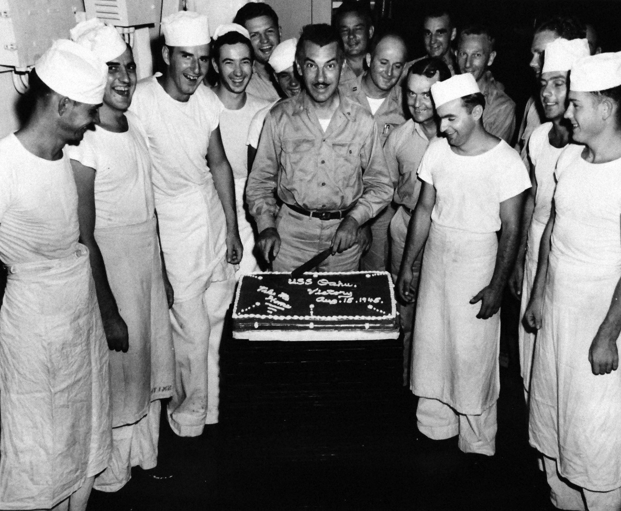 80-G-332797:   V-J Day, August 15, 1945.    USS Oahu (ARG-5), Commanding Officer, Commander A.M. Loker cuts cake in honor of the surrender at Eniwetok, Atoll, Marshall Islands, August 15, 1945. Decommissioned in 1947, she was transferred to the Maritime Administration in July 1963.    Official U.S. Navy photograph, now in the collections of the National Archives. (2015/11/17).   Note, original photograph is extremely curved.  