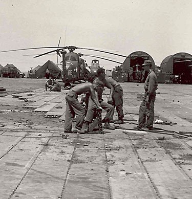 Four Seabees working near an airstrip, with helicopters in the background.
