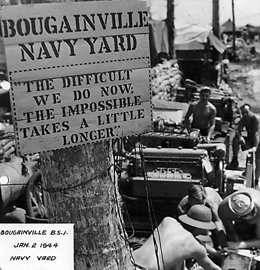 Five Seabees working under a sign that says "Bouganville Navy Yard, The Difficult We Do Now: The Impossible Takes a Little Longer"