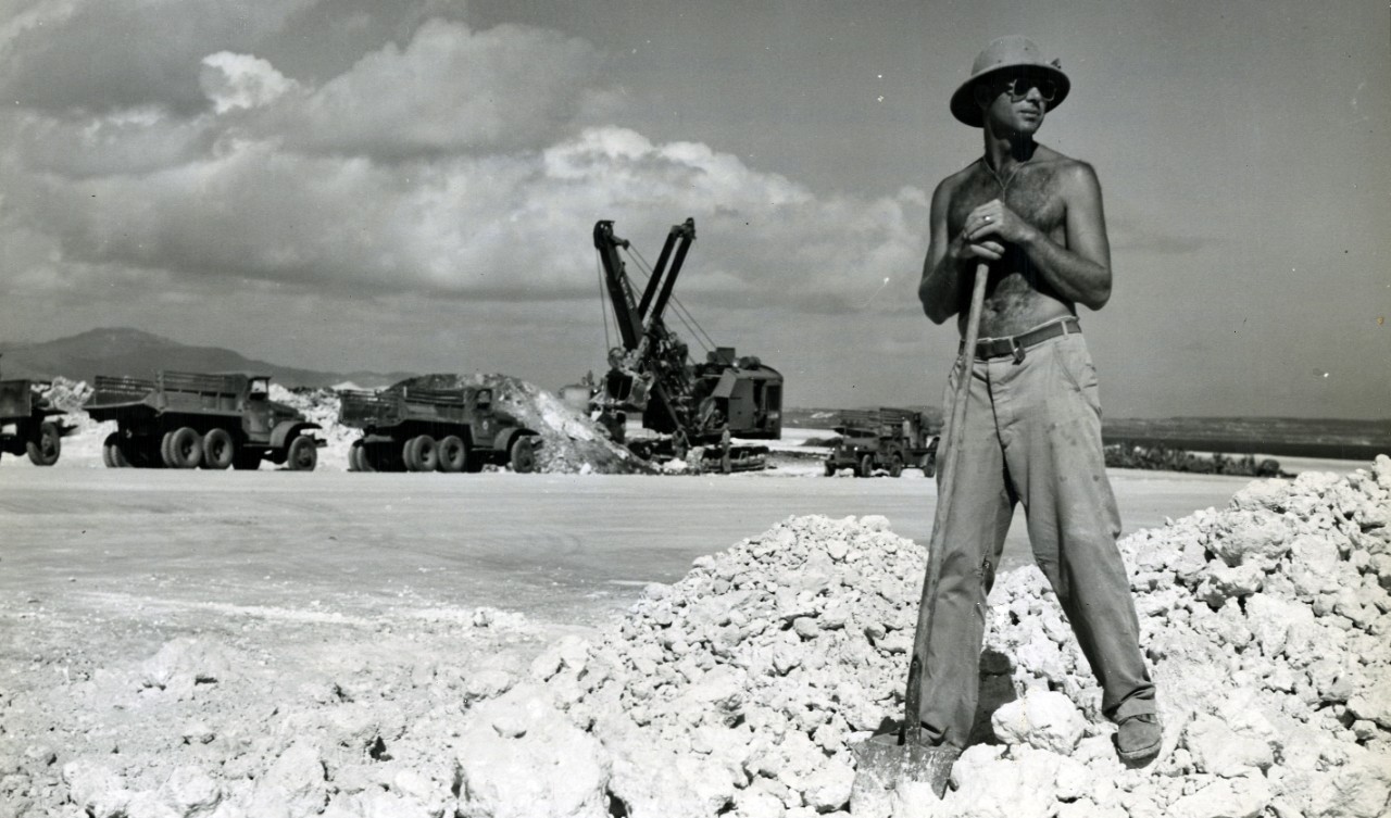 Seabee Vance Shoemate directs truckers unloading coral for runways. The truckers drive at 45 mph. In the background is a fleet of trucks awaiting their turn for leads from the scoop shovel. Working on B-29 bomber base on Tinian.
