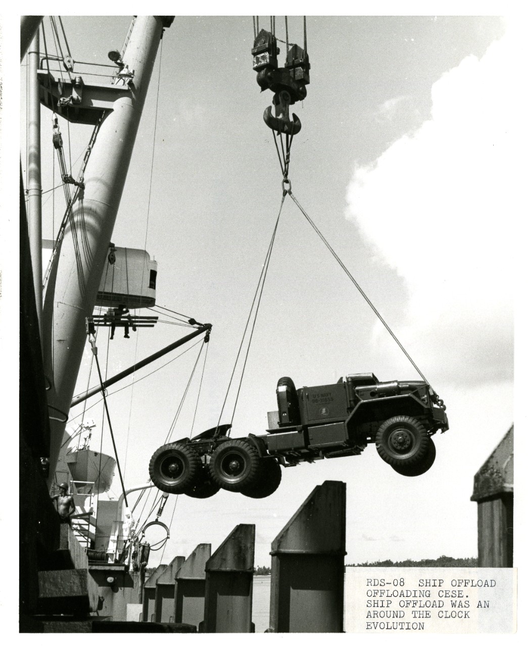 Black and white photograph of a suspended truck in mid-air as a crane offloads a ship at Diego Garcia, British Indian Ocean Territory