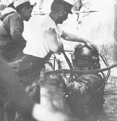 Very old photograph showing a diver in an old-fashioned diving suit being helped into the water by two men. 