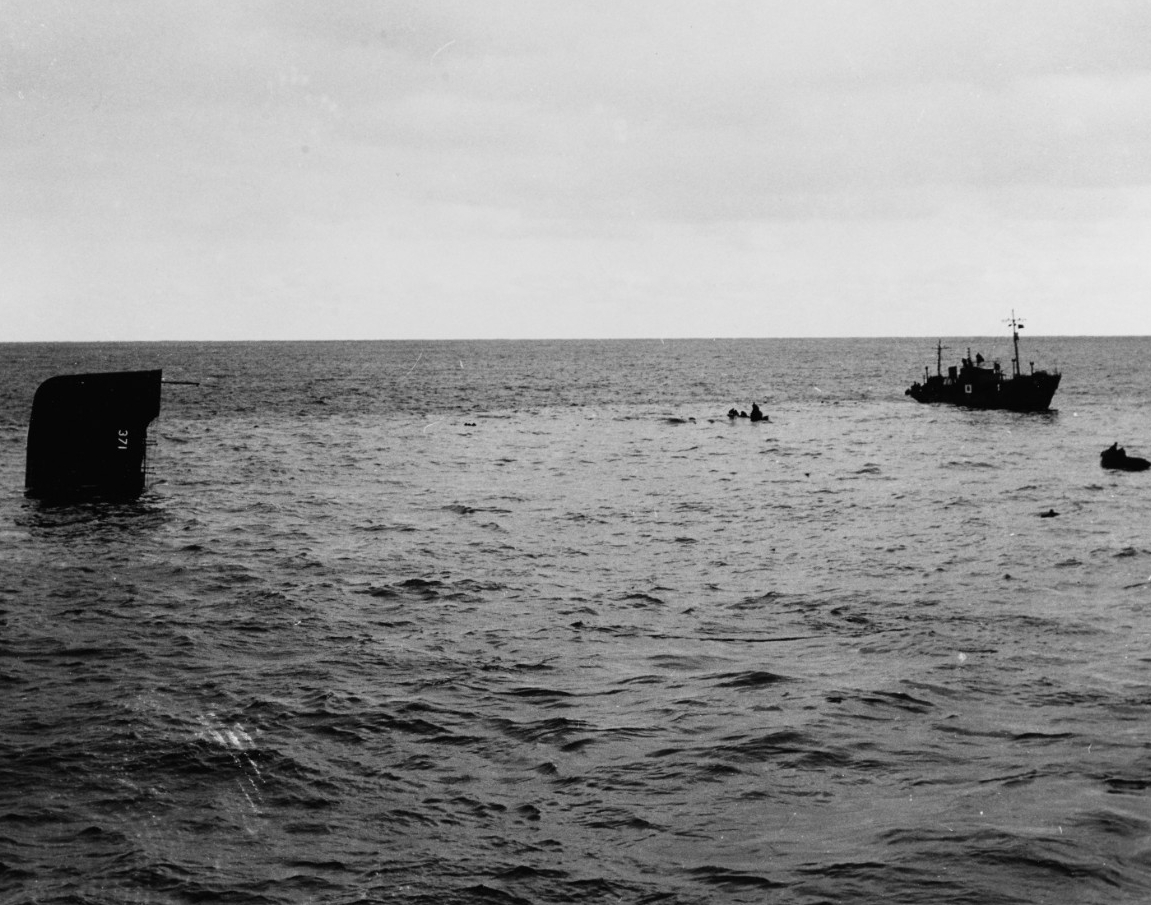 Open sea. A sinking ship, at left, with only a small part of the superstructure visible. On the superstructure is painted the ship’s hull number, 371. At right, background, is a small vessel picking up survivors. 