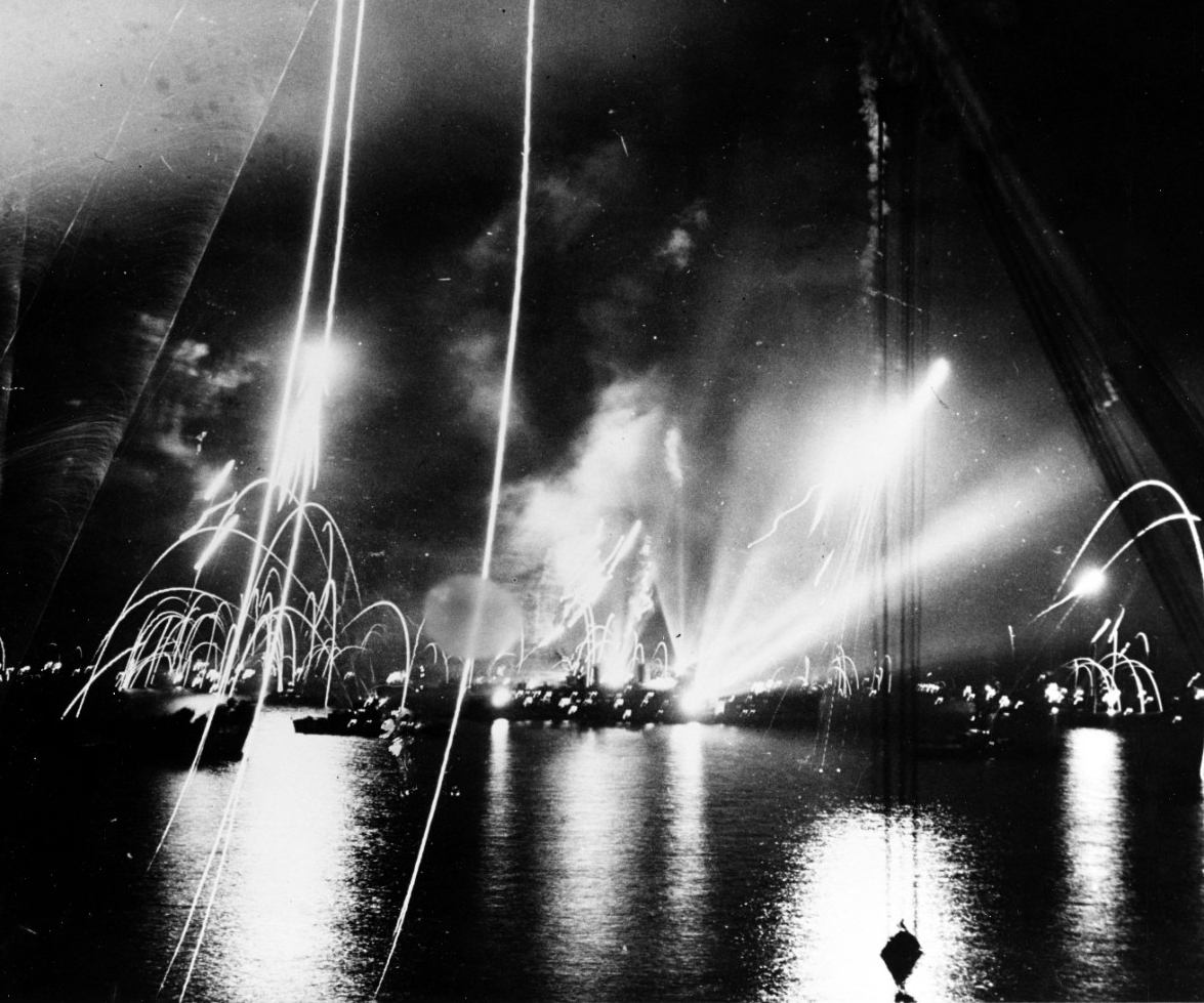 A nighttime photograph showing the arcs of pyrotechnic flares against a dark sky. Reflections of the flares light up the surface of the water.