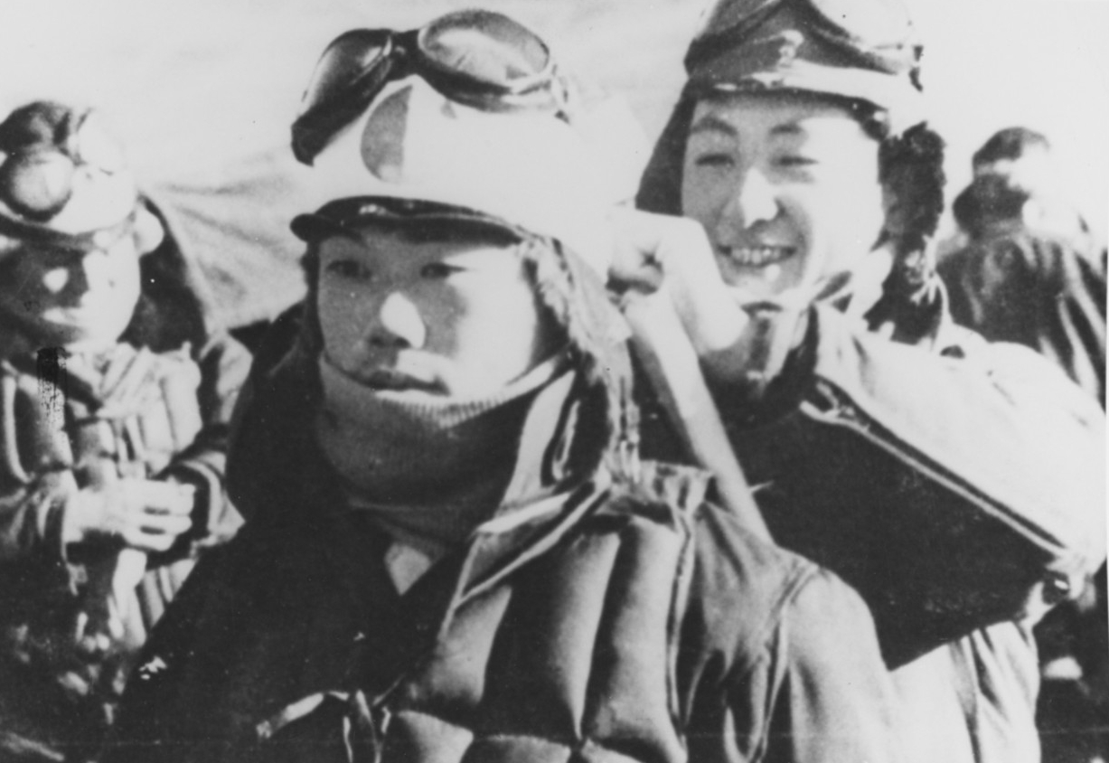 Photo of two Japanese kamikaze pilots preparing for battle. One is wrapping a scarf around the head of the other. 