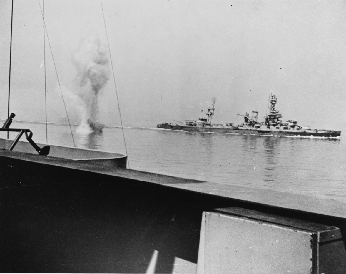 A photograph taken from the deck of one battleship with another battleship in the background with a visible splash between the two ships. 