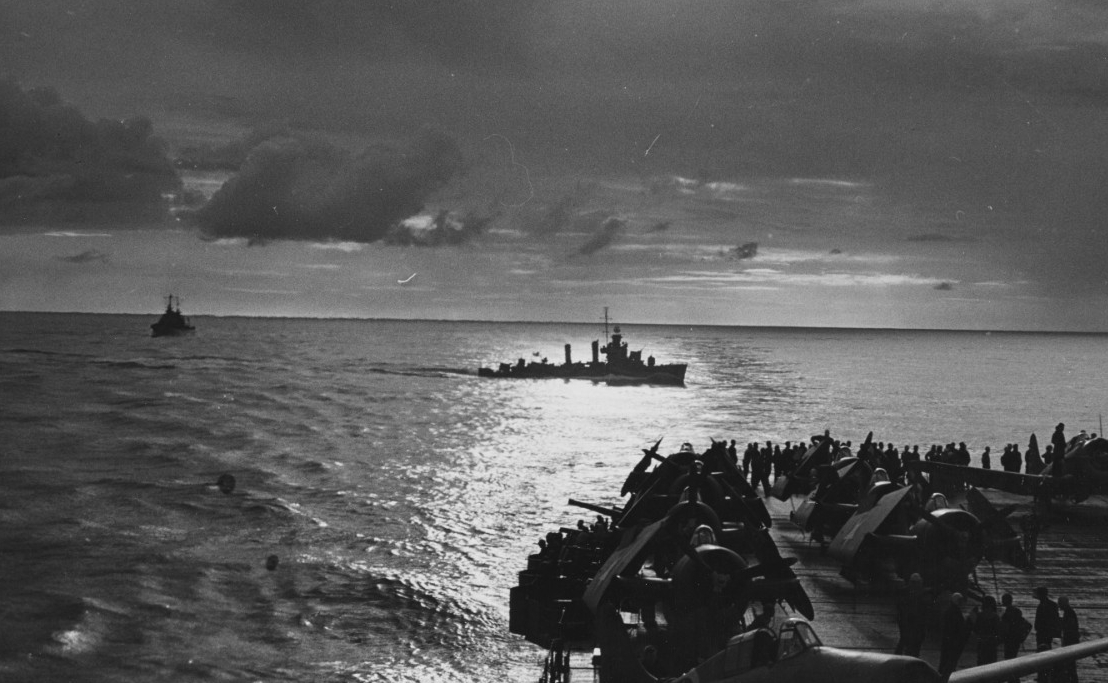 A black-and-white photograph of the view from the deck of an aircraft carrier of a destroyer underway near sunset.