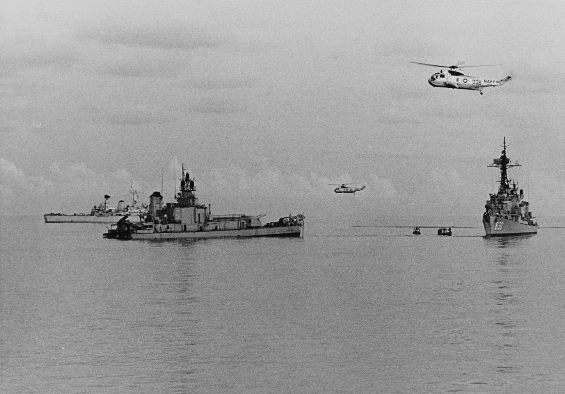 Two helicopters hover over a collection of ships at sea.