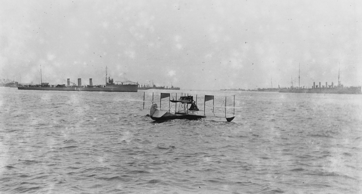 A hydro-aeroplane afloat with a flotilla of destroyers visible in the background.