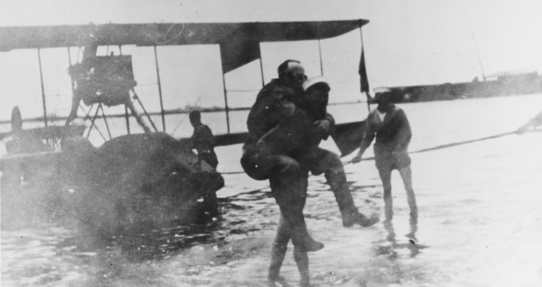 A man is carried on another man’s back from biplane to beach.