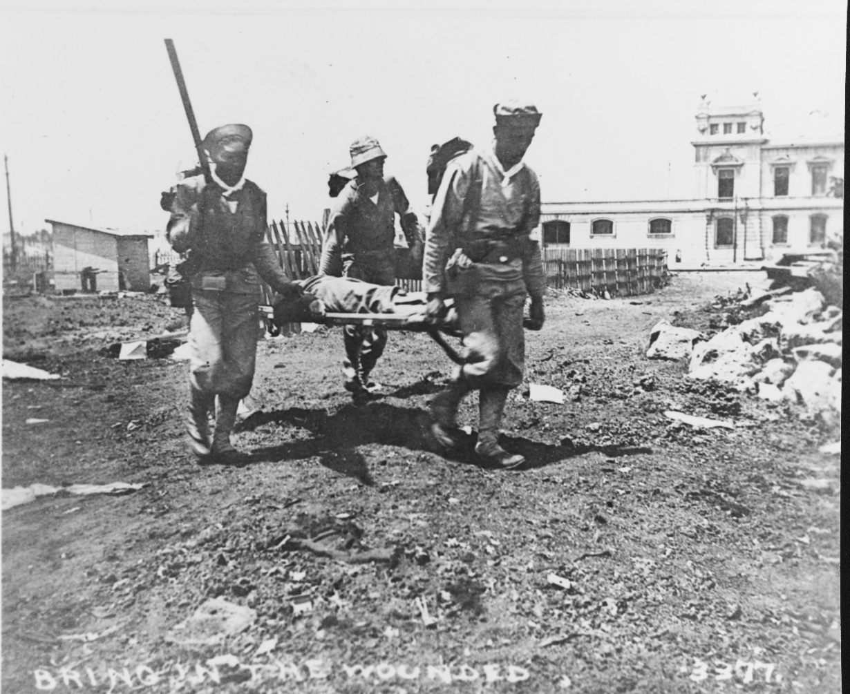 3 men in Navy uniforms carry a stretcher with a wounded person