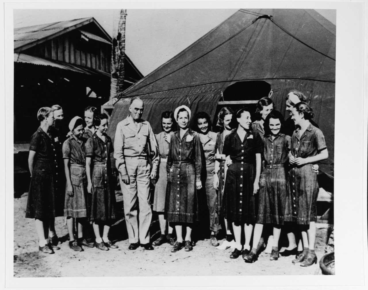 A male flag officer stands next to a group of smiling women in worn-looking dresses outside of a tent