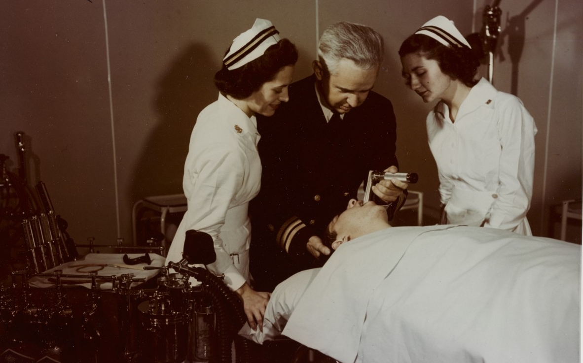 Two women in white nursing uniforms look on as a Navy doctor in uniform inserts a tube into a patients mouth.