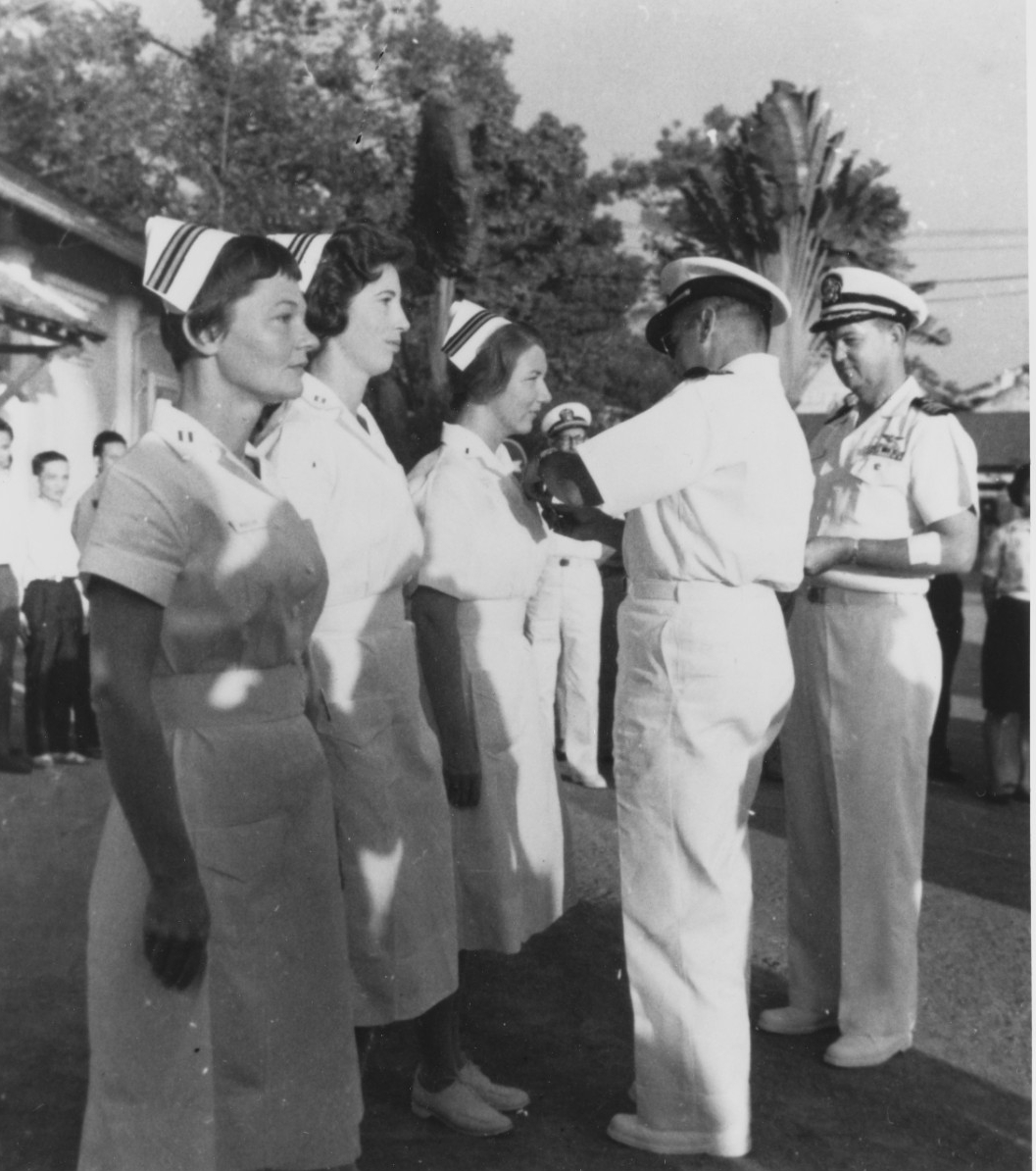 Male officer in white uniform pins an award on a woman in white nursing uniform as two other nurses stand to her right.