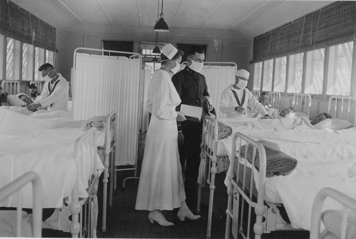 Nurse in all white wears a mask as she talks to a man dressed in black (also wearing a mask). In the background, hospital corpsmen in whites (wearing masks) tend to patients in bed.