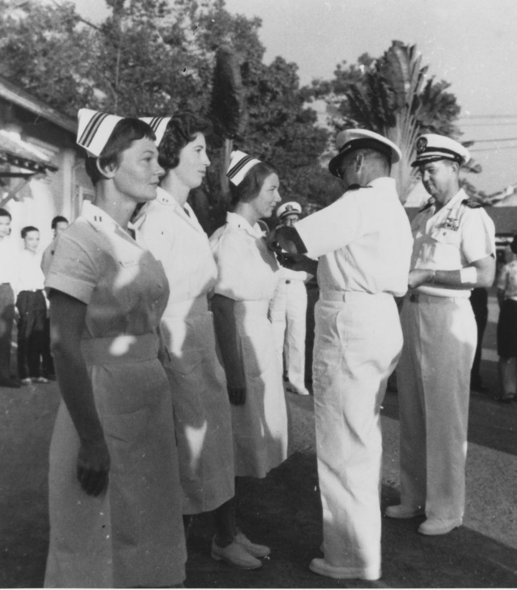 A naval officer pins an award on a nurse standing at attention next to two other nurses to her 