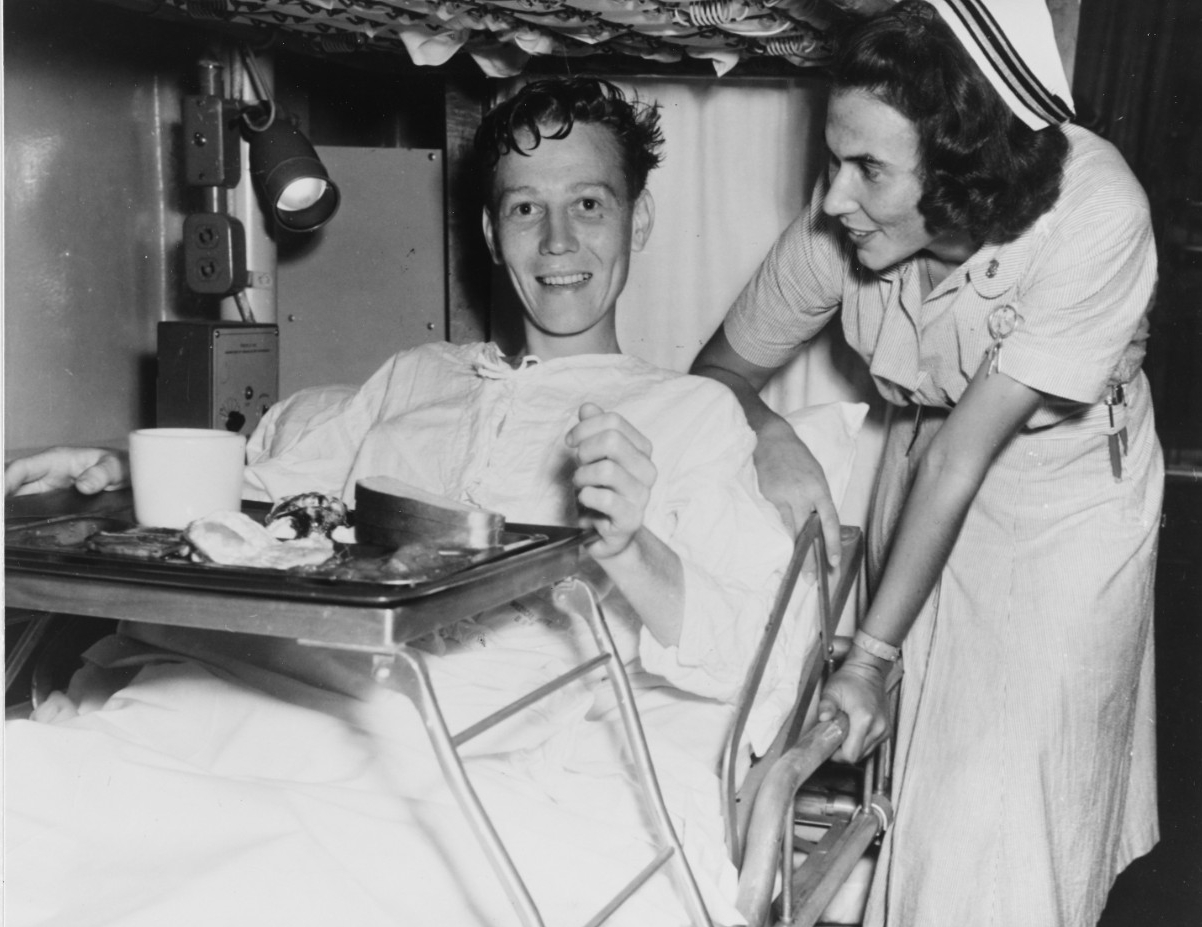 A man in a hospital cot smiles at the camera as a nurse stands next to him