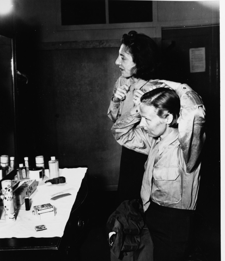 Two women in uniform stand before a mirror and sink combing their hair.