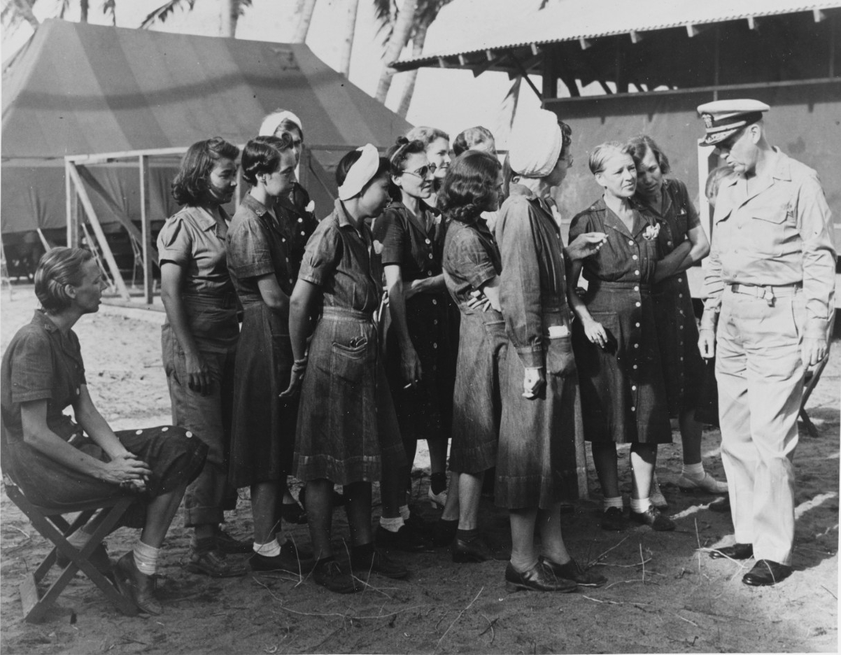 a group of women in denim dresses gather around a naval officer in khaki uniform