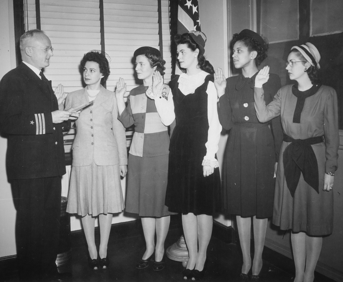 A U.S. Navy officer in uniform holds a paper before him as he faces five young women holding their right hand up