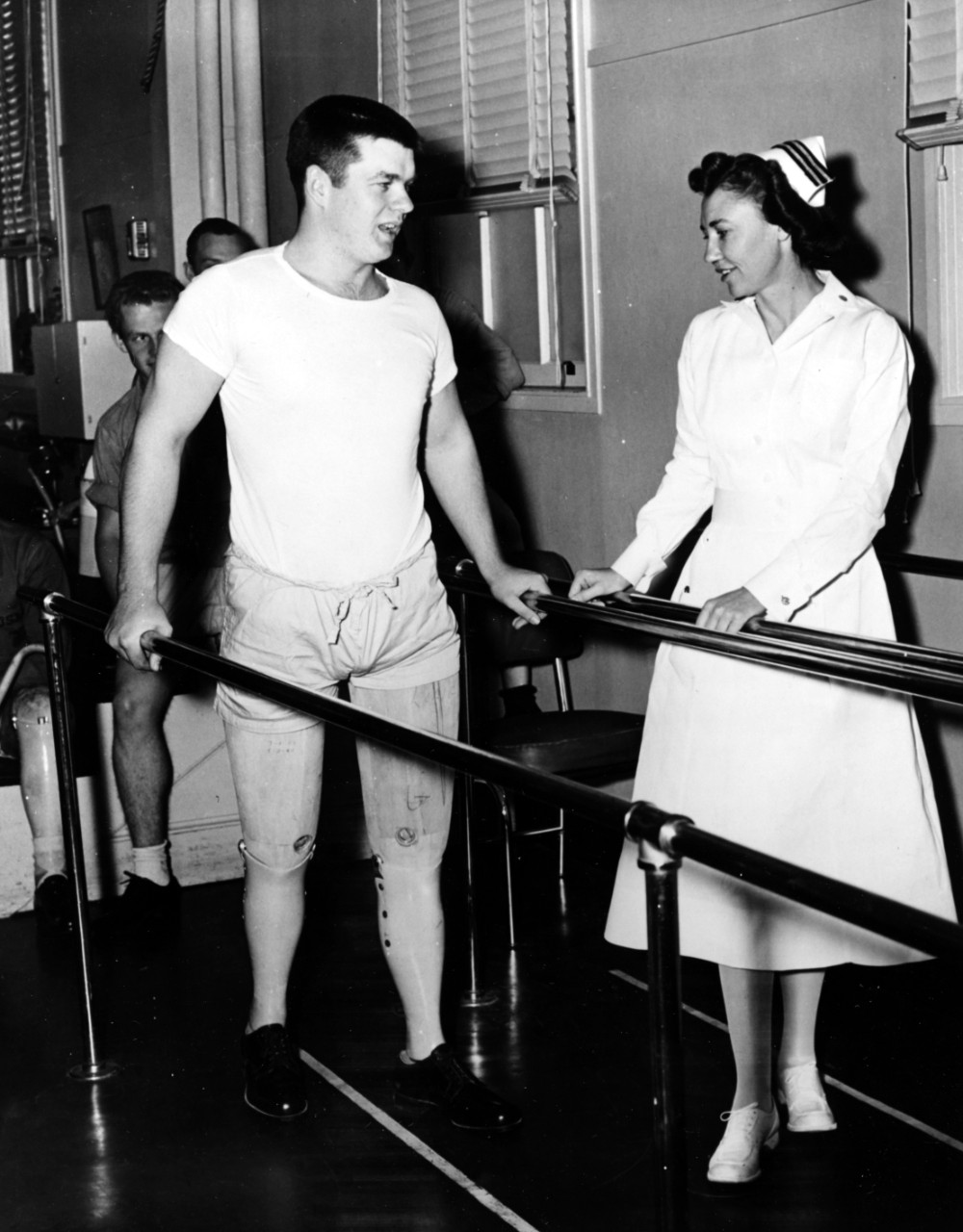A nurse in a white dress and cap talks to a man holding on to handrails.