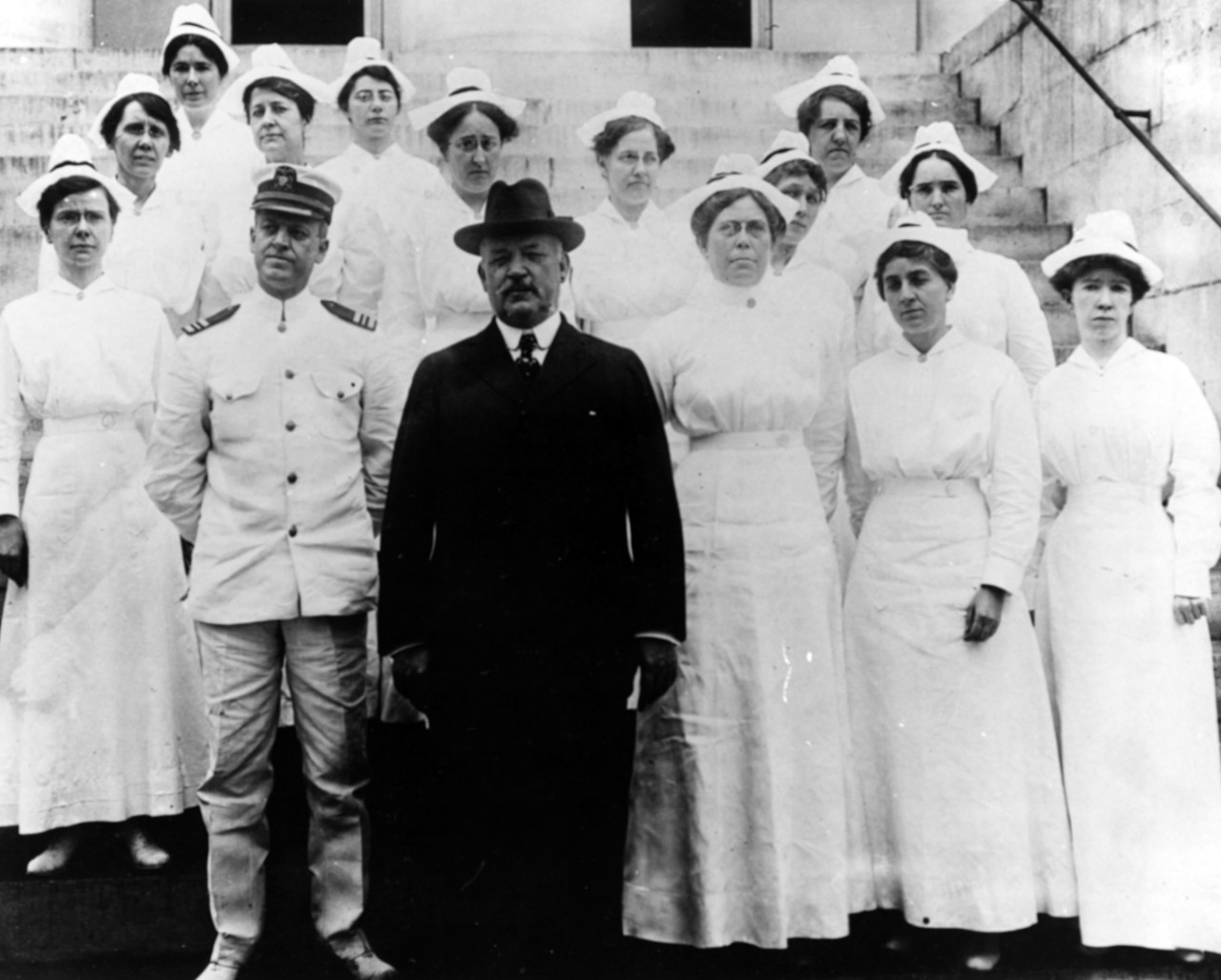 Group of nurses stand on the steps of a building in their white uniform with a male officer and civilian in front.