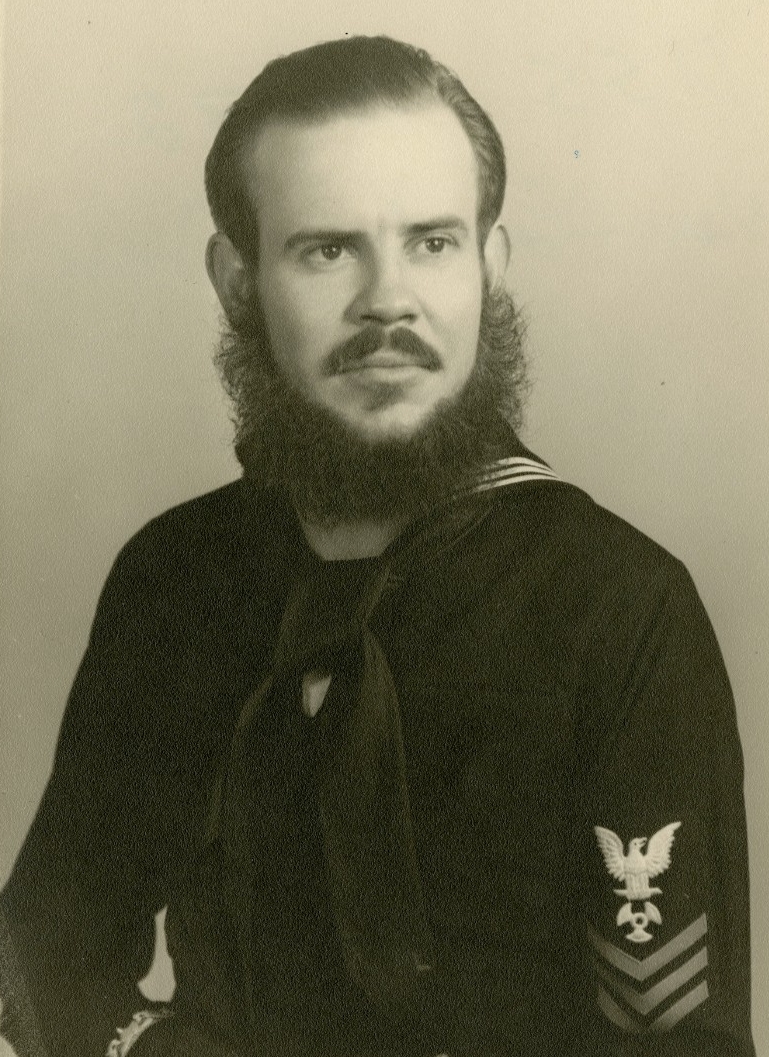 Portrait of a sailor in a Navy dress blue uniform with beard and mustache. The "dolphins" insignia is embroidered on his right sleeve.