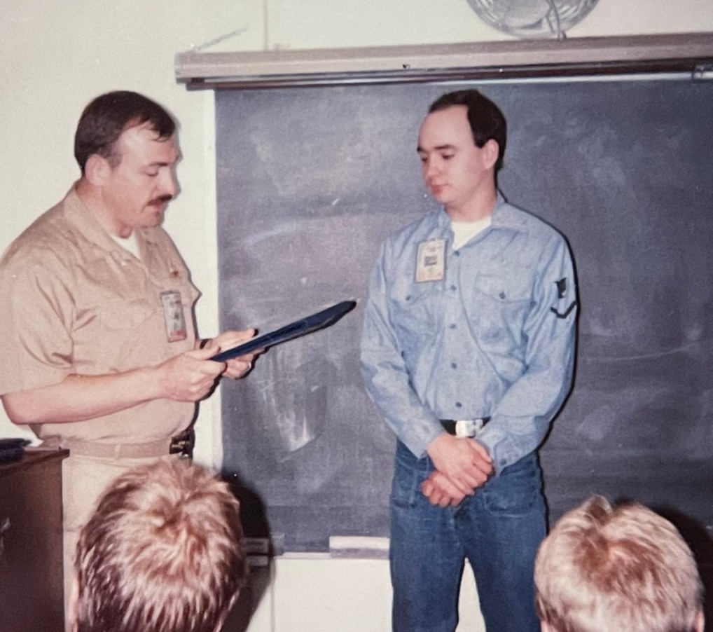 An officer in khakis reads a certificate aloud as an enlisted sailor in dungarees stands by waiting to receive his submarine warfare insignia.
