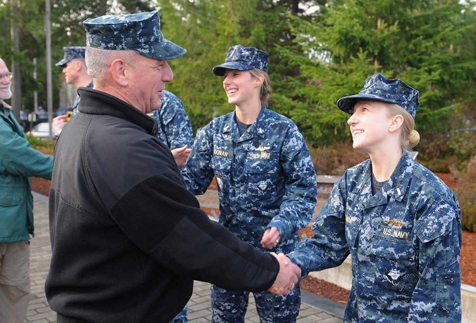A male sailor in uniform shakes the hand of a smiling female officer in uniform.